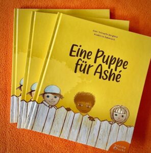Drei der Bücher "Eine Puppe für Ashé" liegen übereinander auf orangenem Untergrund